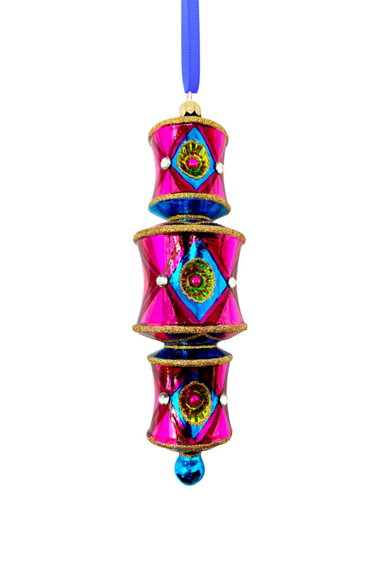 Three-tiered barrel-shaped glass ornament in hot pink deepening to red, with blue diamond reflectors catching flashes of green and pink, faceted crystal accents, gold glitter trim, and a blue glass drop, hung from a blue ribbon.