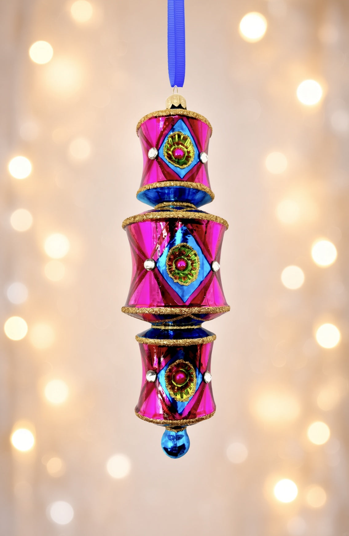 On a soft white holiday lights in the background a three-tiered barrel-shaped glass ornament in hot pink deepening to red, with blue diamond reflectors catching flashes of green and pink, faceted crystal accents, gold glitter trim, and a blue glass drop, hung from a blue ribbon.