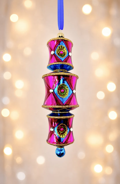 On a soft white holiday lights in the background a three-tiered barrel-shaped glass ornament in hot pink deepening to red, with blue diamond reflectors catching flashes of green and pink, faceted crystal accents, gold glitter trim, and a blue glass drop, hung from a blue ribbon.