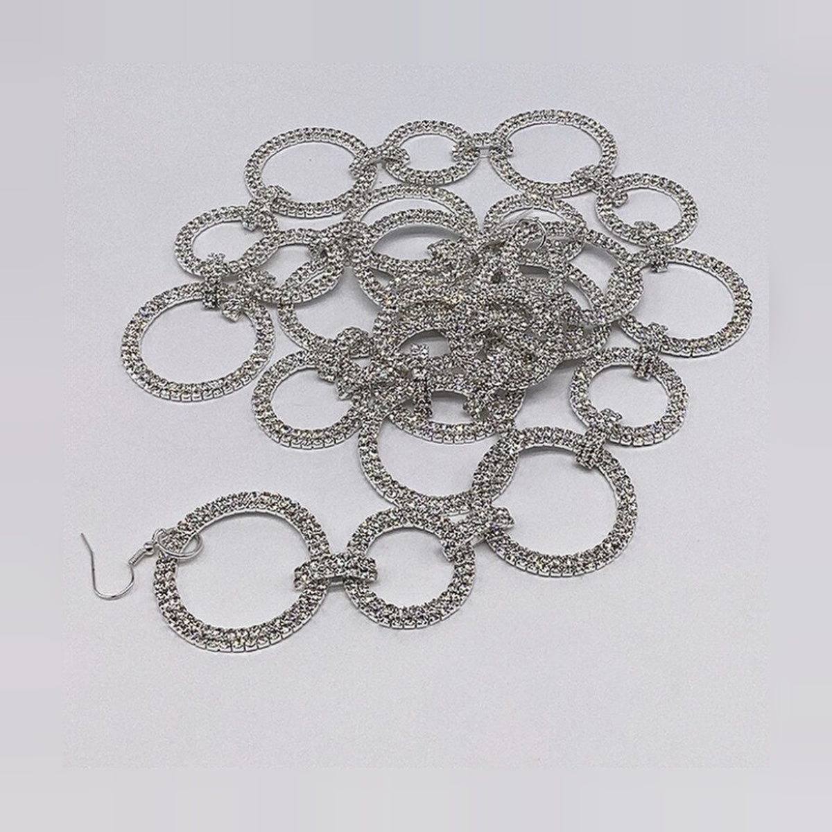 Silver garland coiled loosely on a pale gray surface, showing alternating small and large open rings, each paved in a double row of clear crystals and linked by short crystal-covered bars, with a silver wire hook at one end.