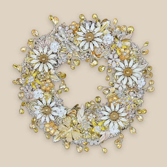 Champagne gold jeweled wreath with white enamel daisies, faux pearl-centered blooms, a faux pearl butterfly, rhinestone florals, gold leaf accents and cascading champagne teardrop rhinestones on a warm stone background.