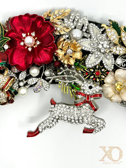 Close-up of jeweled Christmas wreath featuring a rhinestone reindeer with red bow, red enamel flower with pearl accent center, and sparkling holiday details