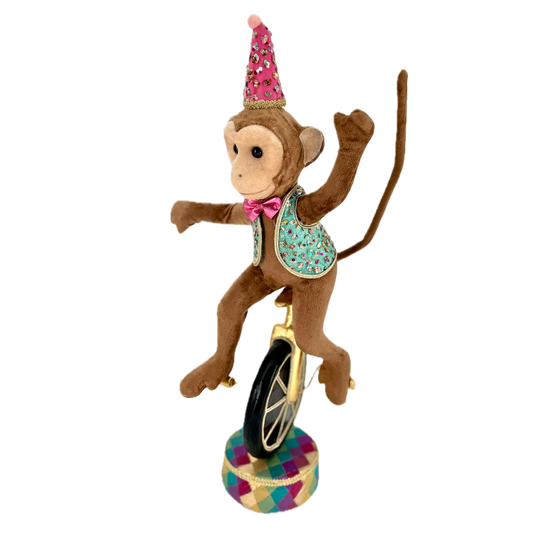 Brown monkey figure with a tan face riding a unicycle with a black wheel and gold frame, arms outstretched and long tail raised upward behind it. It wears a sparkling pink cone-shaped party hat, a pink bow tie and a blue vest dotted with colorful sequins and gold trim, on a round pedestal with a raspberry, teal, gold and purple harlequin pattern.