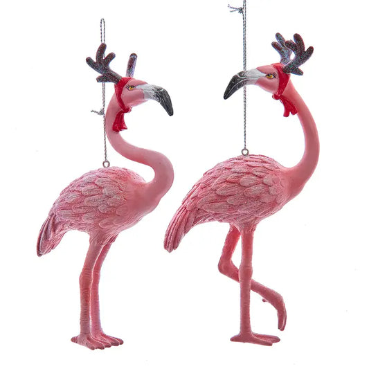Two glittered pink flamingo Christmas ornaments wearing glittered reindeer antlers and red scarves, one standing and one posed on one leg.