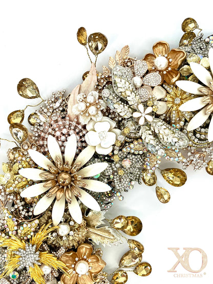 A close-up of a jeweled wreath featuring embelished jeweled dragonfly, white enamel daisy brooches, faux pearl-center florals, champagne rhinestones, gold leaf accents, and faceted amber teardrop rhinestones