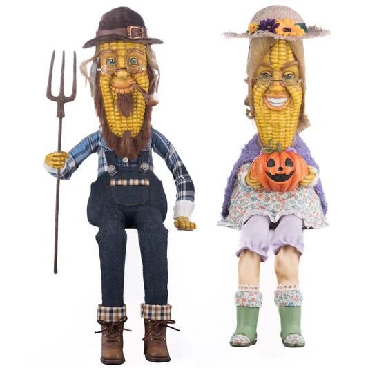 Mr. and Mrs. Cobb lanky legs folk art figurine set — corn cob-faced couple with poseable legs, he in denim overalls with a pitchfork, she in a floral dress with a jack-o-lantern, on a white background.