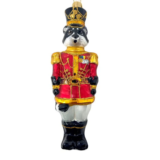 Raccoon nutcracker soldier ornament in a red and gold ceremonial military coat with gold epaulettes and a striped ribbon medal, black tall guard's hat with gold trim, white trousers, black boots, and the raccoon's natural dark eye-mask markings, shown against a white background.